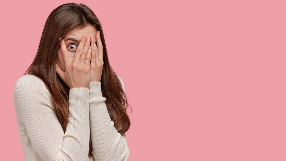 photo scared woman covers face with both hands looks through fingers peeks with fear has dark hair dressed comfortable jumper