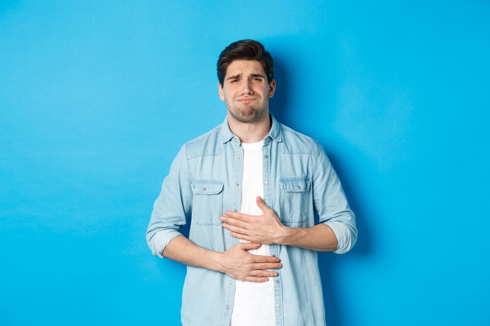 man holding hands belly grimacing from pain complaining stomach ache standing against blue background 1258 66410
