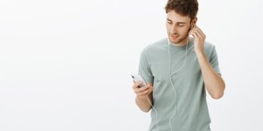 portrait-relaxed-good-looking-european-male-model-with-bristle-holding-hand-earphone-looking-smartphone-screen-while-enjoying-great-sound-music-earbuds_176420-24992
