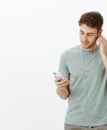 portrait-relaxed-good-looking-european-male-model-with-bristle-holding-hand-earphone-looking-smartphone-screen-while-enjoying-great-sound-music-earbuds_176420-24992