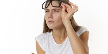 Picture of focused serious young Caucasian female teacher with textbook, taking off glasses and narrowing eyes, trying to see something clearly. Student girl in eyewear posing with diary in studio